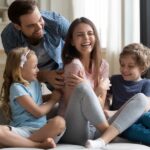 A family of four is laughing together on a couch in a brightly lit living room.
