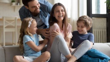 A family of four is laughing together on a couch in a brightly lit living room.