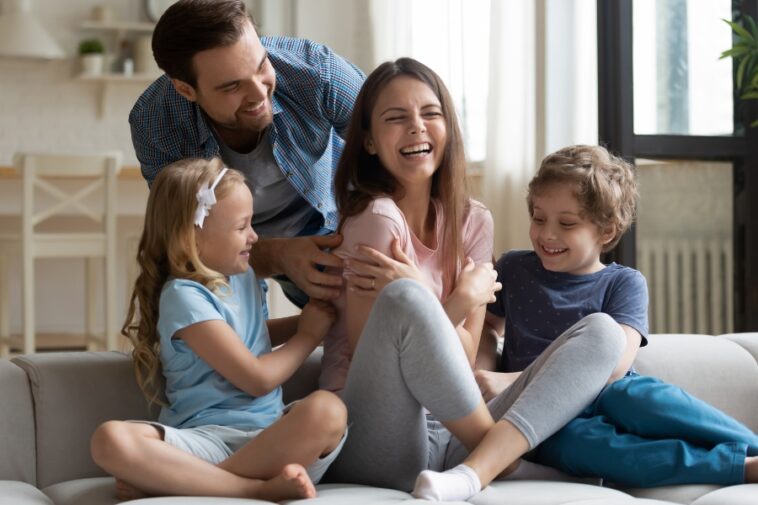 A family of four is laughing together on a couch in a brightly lit living room.