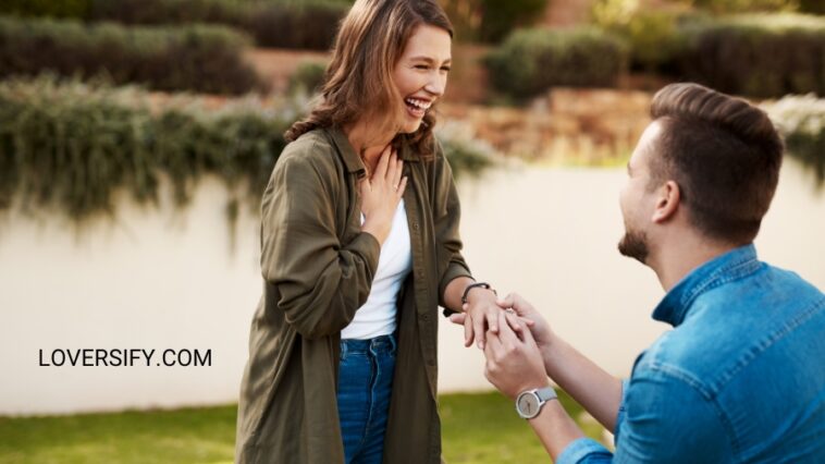 Outdoors, a man kneels, holding a woman's hand, as she laughs with her hand on her chest.