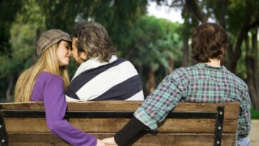 In a park setting, a couple sits on a bench, faces close, while a person in a plaid shirt sits on the other side with their back to the viewer; the couple's hands are intertwined behind the bench.
