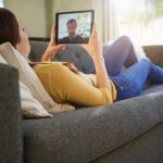 Woman lying on a gray couch, holding a tablet with a man visible on the screen.