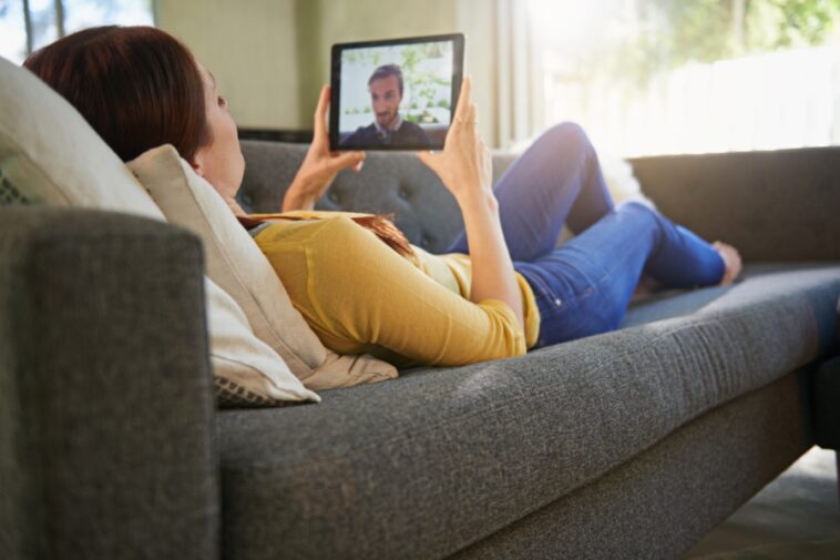 Woman lying on a gray couch, holding a tablet with a man visible on the screen.
