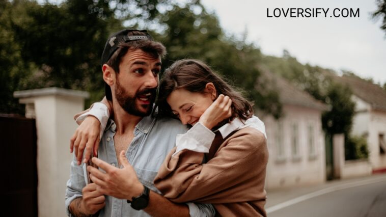 A man and woman embrace on a street with houses and trees in the background.