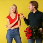 Against a yellow backdrop, a man with roses and a ring box faces a woman who is holding up her hand in a "no" gesture.