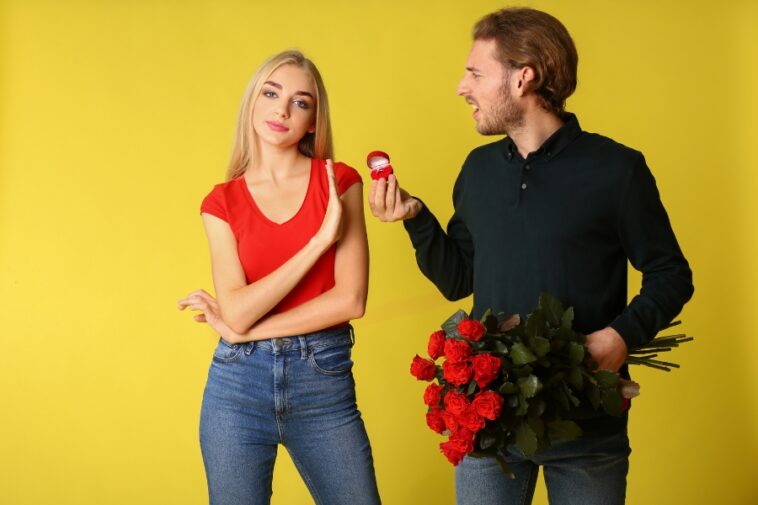 Against a yellow backdrop, a man with roses and a ring box faces a woman who is holding up her hand in a "no" gesture.