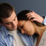 A man in a blue striped shirt comforts a woman in a white shirt, his hand on her head.