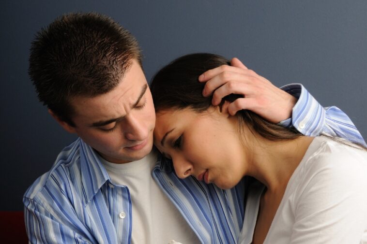 A man in a blue striped shirt comforts a woman in a white shirt, his hand on her head.