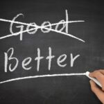 Chalkboard with the word "Good" crossed out and "Better" underlined by a hand holding chalk.