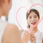 A smiling woman draws a pink heart on a mirror, looking at her reflection.