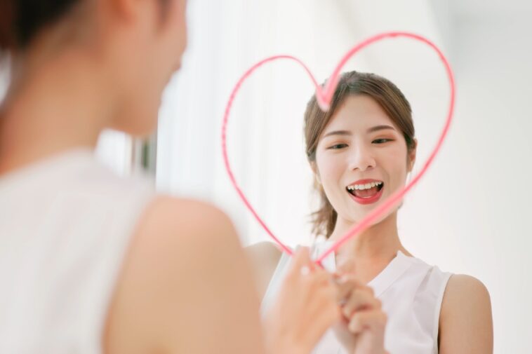 A smiling woman draws a pink heart on a mirror, looking at her reflection.