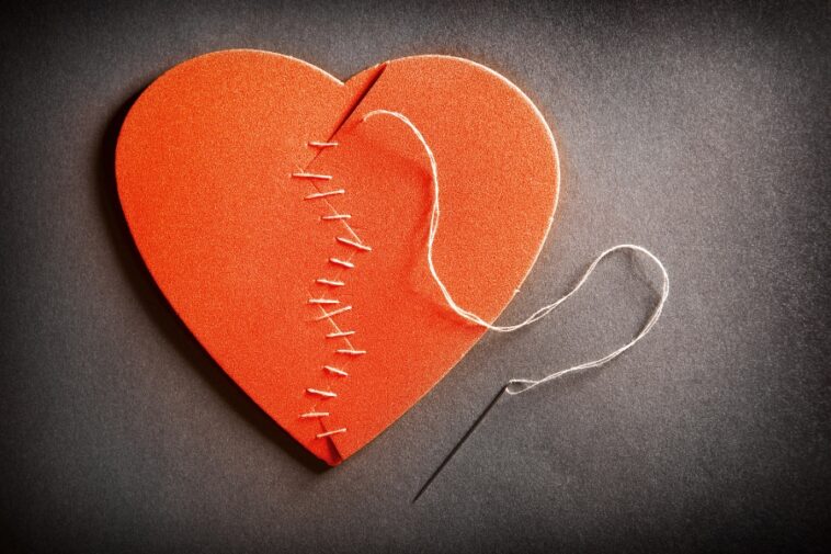 An orange heart shape is stitched together with white thread on a gray surface, a needle lies nearby.