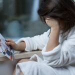 Woman in a white robe sits by a window, holding a torn photograph of a couple.