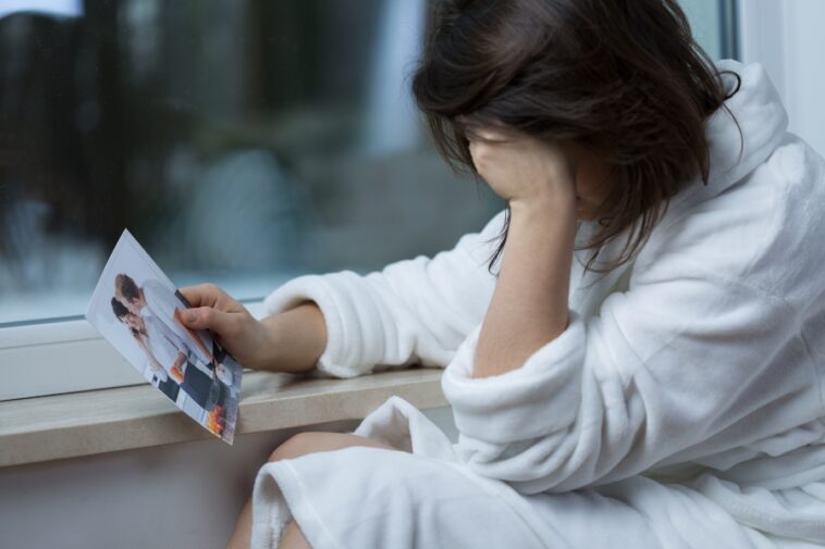 Woman in a white robe sits by a window, holding a torn photograph of a couple.