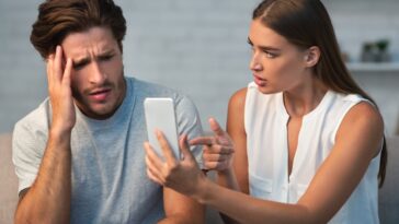 A man and woman are looking at a smartphone, with the woman pointing at the screen and the man holding his head with a concerned expression.