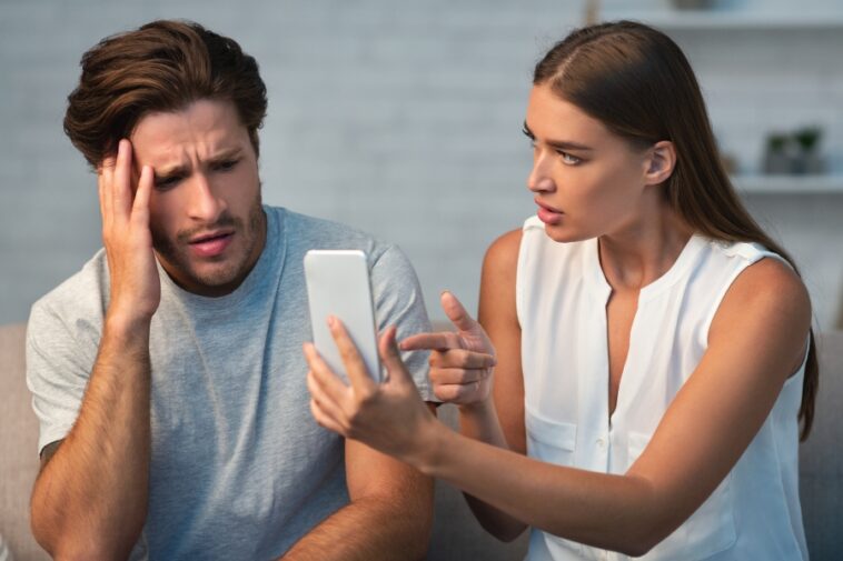 A man and woman are looking at a smartphone, with the woman pointing at the screen and the man holding his head with a concerned expression.