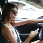 Inside a car, a woman with sunglasses and a seatbelt is holding a phone while one hand is on the steering wheel.