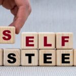 A hand places a wooden block with a red "S" on top of other blocks to spell "SELF ESTEEM".