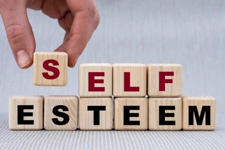 A hand places a wooden block with a red "S" on top of other blocks to spell "SELF ESTEEM".