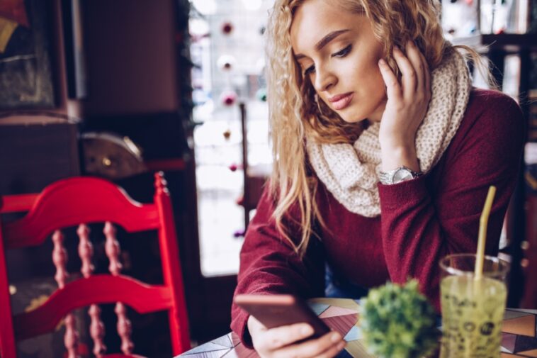 A young woman with blonde curly hair, wearing a scarf and sweater, looks at her phone in a cafe setting.