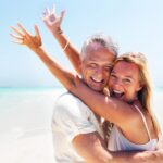 A smiling couple embraces on a sunny beach, with the woman's arms raised in the air.