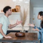 In a kitchen setting, a man and a woman stand facing each other with raised hands, appearing to be in a heated discussion.