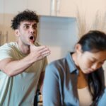 In a kitchen setting, a man with curly hair gestures emphatically while appearing to shout at a woman with her head bowed and hand on her forehead.