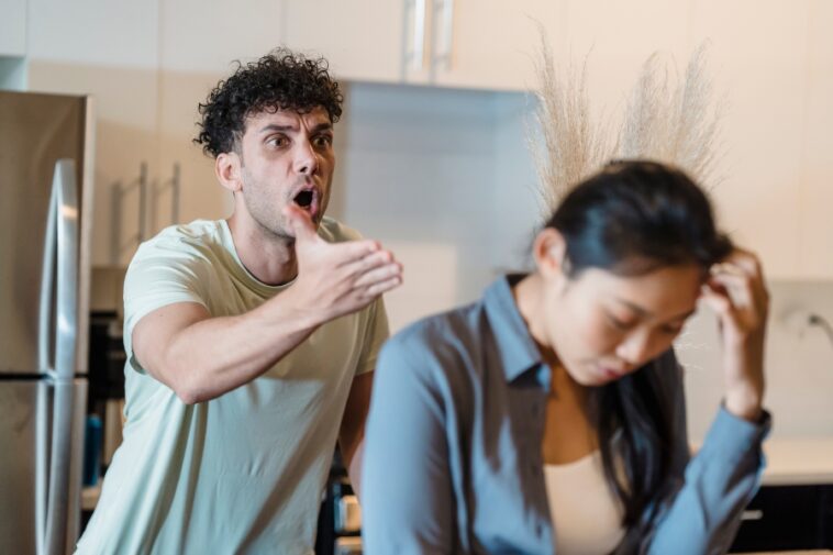 In a kitchen setting, a man with curly hair gestures emphatically while appearing to shout at a woman with her head bowed and hand on her forehead.