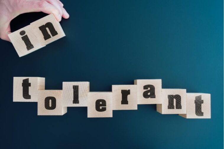Hand placing a wooden block with "in" on top of blocks spelling "tolerant" against a blue background.