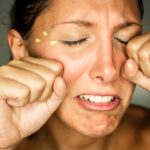 Close-up of a person with closed eyes, clenching their fists and teeth, with small yellow shapes near one eye.