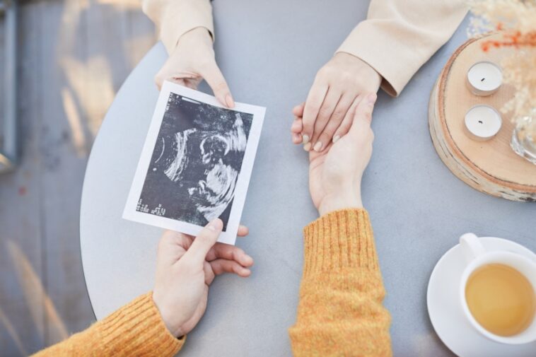 Overhead view of two people holding hands at a table; one person holds an ultrasound image. A cup of tea and small candles are also on the table.