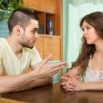 A man and a woman sit at a table facing each other, seemingly in conversation. The man gestures with his hand, while the woman looks at him with her hands clasped.