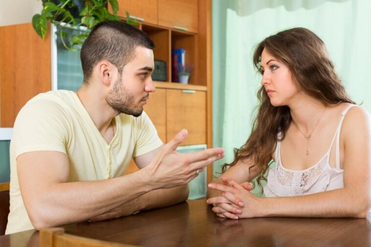 A man and a woman sit at a table facing each other, seemingly in conversation. The man gestures with his hand, while the woman looks at him with her hands clasped.