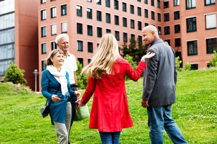 Four people stand on a grassy hill in front of a brick building. A woman in a red coat touches the shoulder of a man in a gray suit. An older couple stands nearby, smiling.