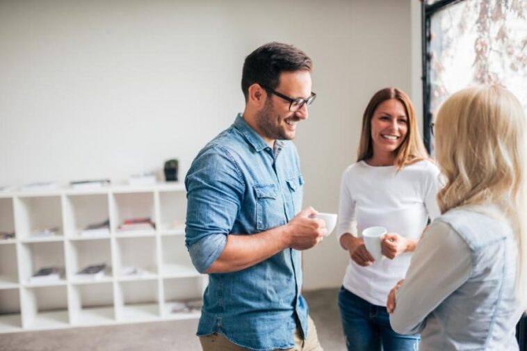 Three smiling people in a bright room are having a conversation while holding mugs.