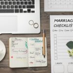 Overhead shot of a desk with a laptop, wedding rings, coffee, notebook, pen, checklist, calendar, and flowers.