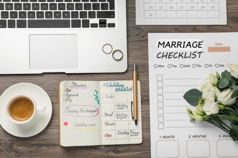 Overhead shot of a desk with a laptop, wedding rings, coffee, notebook, pen, checklist, calendar, and flowers.