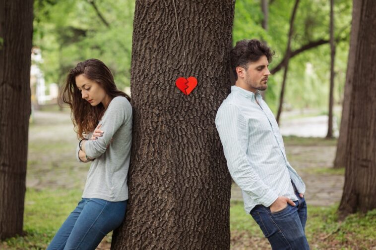 Outdoors, a woman and a man stand on opposite sides of a tree with a broken heart symbol on it.