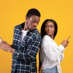 On a yellow backdrop, a man and woman stand back-to-back, each holding a smartphone. The man glances at the woman's phone, while she looks away with a suspicious expression.