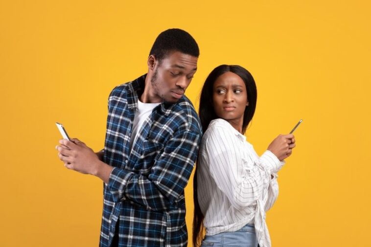 On a yellow backdrop, a man and woman stand back-to-back, each holding a smartphone. The man glances at the woman's phone, while she looks away with a suspicious expression.