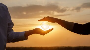 Against a sunset backdrop, one person holds a glowing, geometric heart in their palm while another person's hand hovers protectively above.