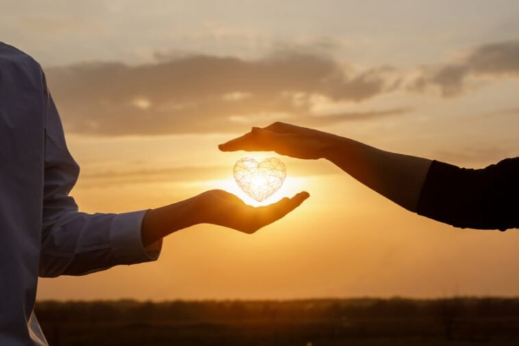 Against a sunset backdrop, one person holds a glowing, geometric heart in their palm while another person's hand hovers protectively above.