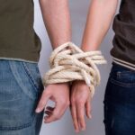 Two people stand side-by-side with their wrists bound together by thick rope.