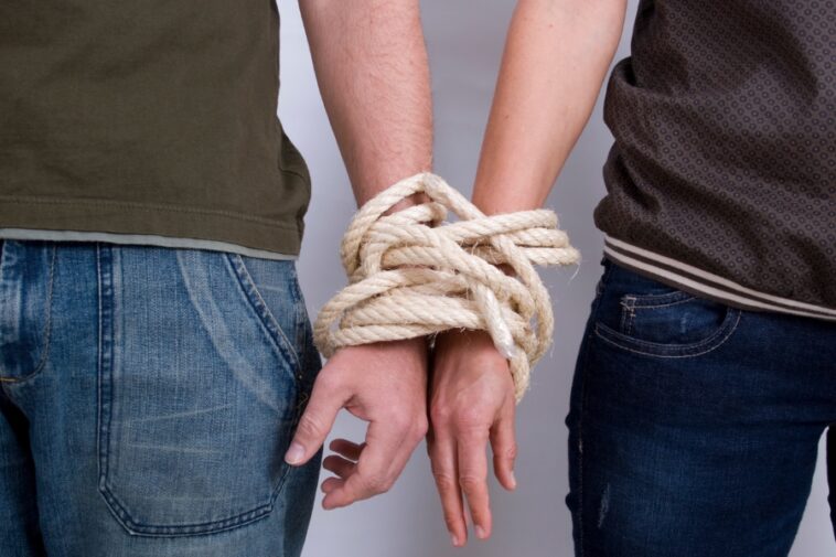 Two people stand side-by-side with their wrists bound together by thick rope.