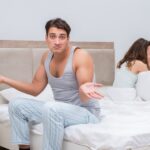 In a bedroom, a man in pajamas sits on the edge of the bed with a questioning expression, arms outstretched. Behind him, a woman sits with her face in her hands.