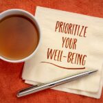 Overhead shot of a cup of tea, a stack of napkins printed with "Prioritize Your Well-Being," and a pen, all on an orange textured surface.