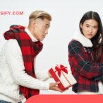 A man offers a gift to a woman who looks displeased. He wears a white sweater and red plaid scarf, while she wears a red plaid jacket with a white collar. A red and white gift box is held out to her.
