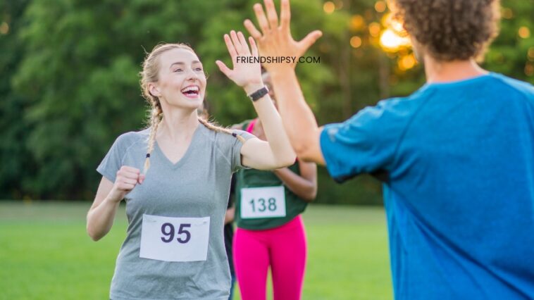 A smiling woman with braids, wearing a race number, gives a high five to a person in a blue shirt; other runners are nearby on a grassy field.