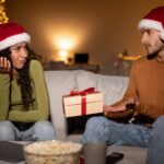 A man in a Santa hat offers a small gift to a woman in a Santa hat who looks unimpressed. They are sitting on a couch in a room lit by string lights.