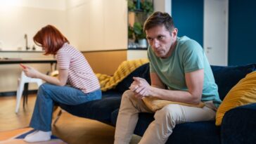 A man and woman sit apart on a blue couch, the woman looks at her phone, and the man looks away.